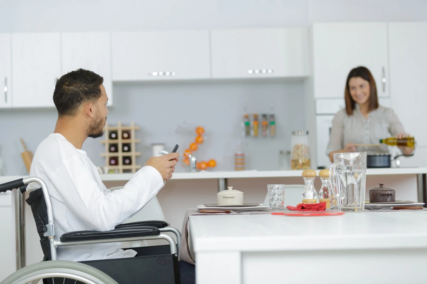 Disabled man in wheelchair sitting at kitchen bench talking to carer