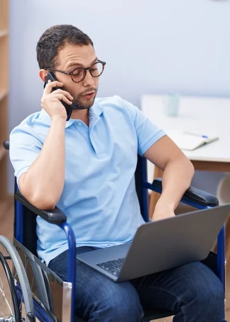 Disabled man in wheelchair with laptop, using phone to make a call