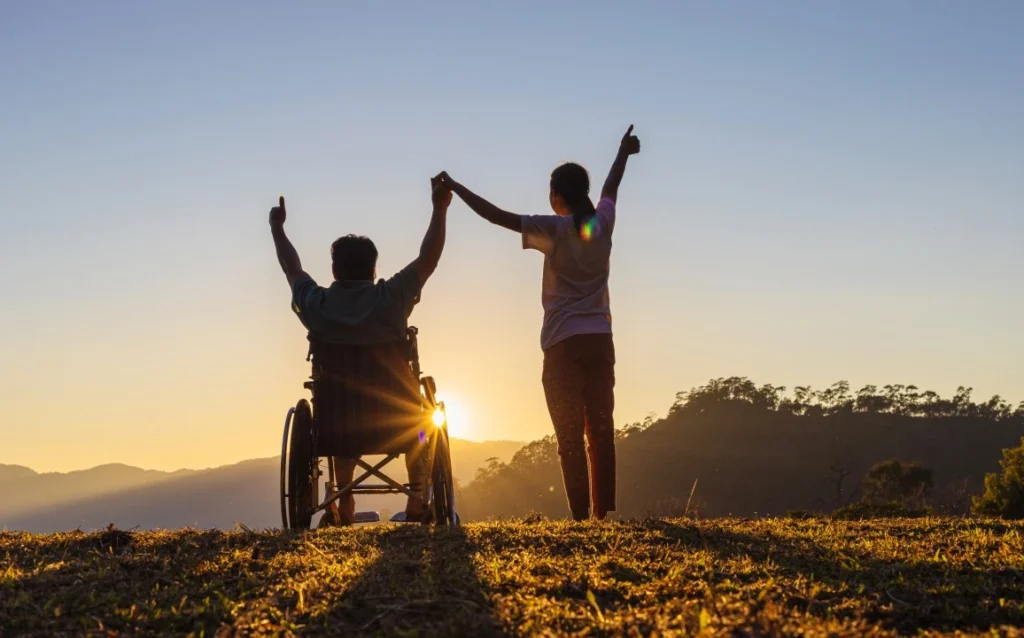 Two people, one person in wheelchair, standing outside holding hands at sunset